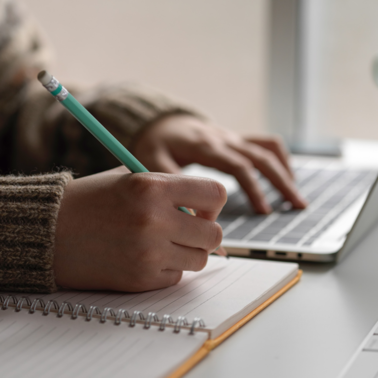 An image of someone typing on a laptop with a notebook beside them as if studying