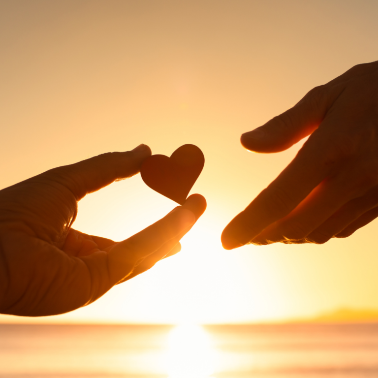 An image of a sunset in the background and 2 hands handing each other a heart shaped pebble to illustrate being kind and sharing kindness