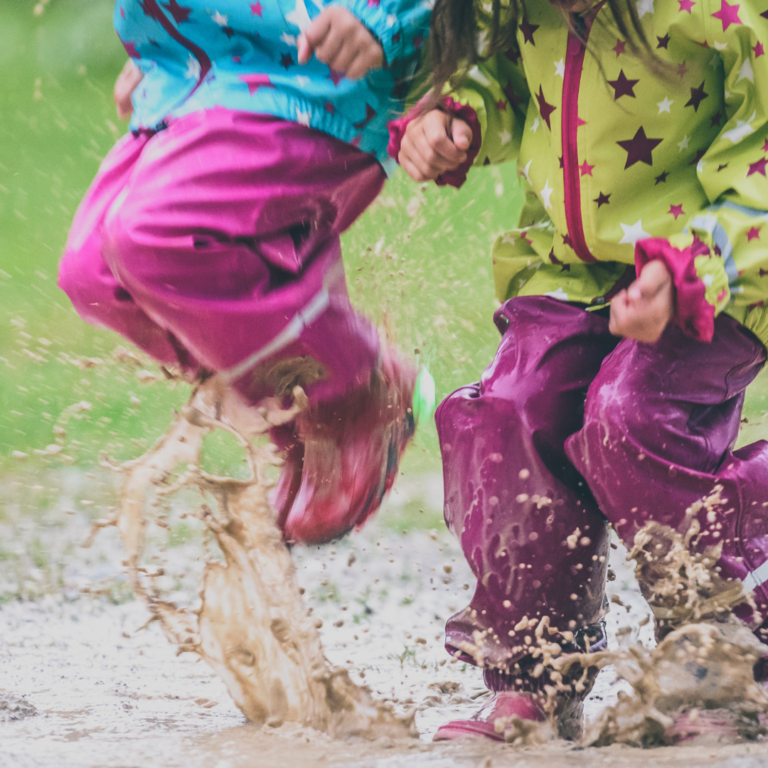 An image of 2 children wearing wellies and raincoats and jumping in mud